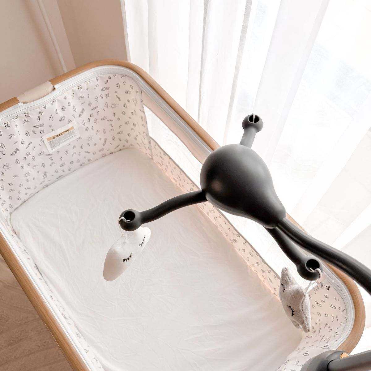 Baby Bassinet & Bedside Co-Sleeper with beige fabric and light wood-grain frame featuring a white fitted sheet and hanging toys