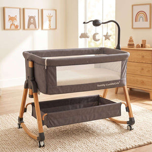 Baby Bassinet & Bedside Co-Sleeper in dark grey fabric with wood-grain steel frame, mesh sides, hanging mobile with stars and clouds, and under-bassinet storage basket