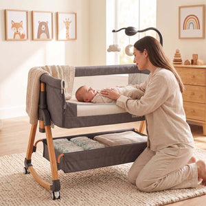 Baby Bassinet & Bedside Co-Sleeper in grey fabric with wooden legs, mesh sides, storage basket underneath, and mobile toy, with mother interacting with baby