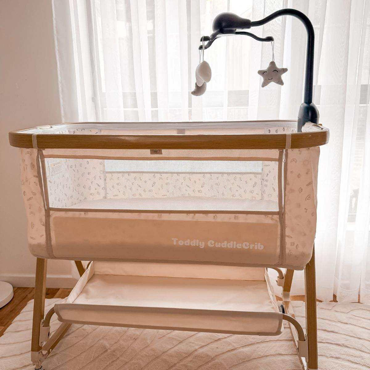 Toddly CuddleCrib V2 Baby Bassinet & Bedside Co-Sleeper in beige linen-cotton blend with wood grain steel frame, mesh sides, under-bassinet storage, and attached black mobile arm with hanging toys