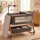 Toddly CuddleCrib V2 Baby Bassinet & Bedside Co-Sleeper in grey fabric with woodgrain steel legs, mesh sides, rocking base, wheels, and a removable mattress beside hanging sensory toys