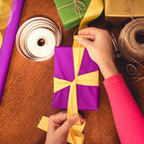 Hands wrapping a purple gift box with yellow ribbon and other wrapping supplies on wooden table for gift wrap service